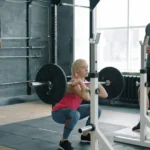 Woman doing barbell squat under trainer supervision in gym