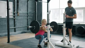 Woman doing barbell squat under trainer supervision in gym